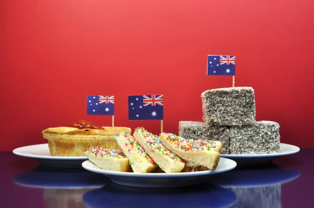 Traditional Australian food - meat pie and sauce, lamingtons and fairy bread - with flag. Australia Day January 26, or Anzac Day celebrate with tradional Aussie tucker food such as lamingtons, meat pies and tomato sauce, and yummy fairy bread.