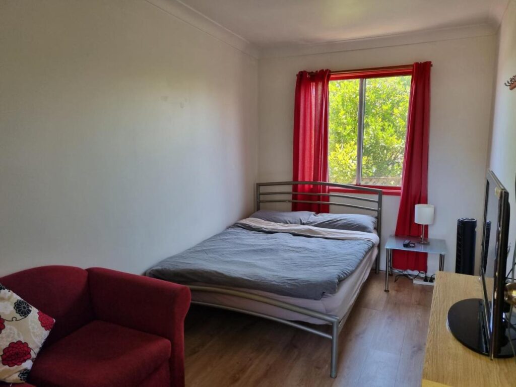 A private room with large wondow with red cyrtains and a red chair next to the bed at Beachside Backpackers