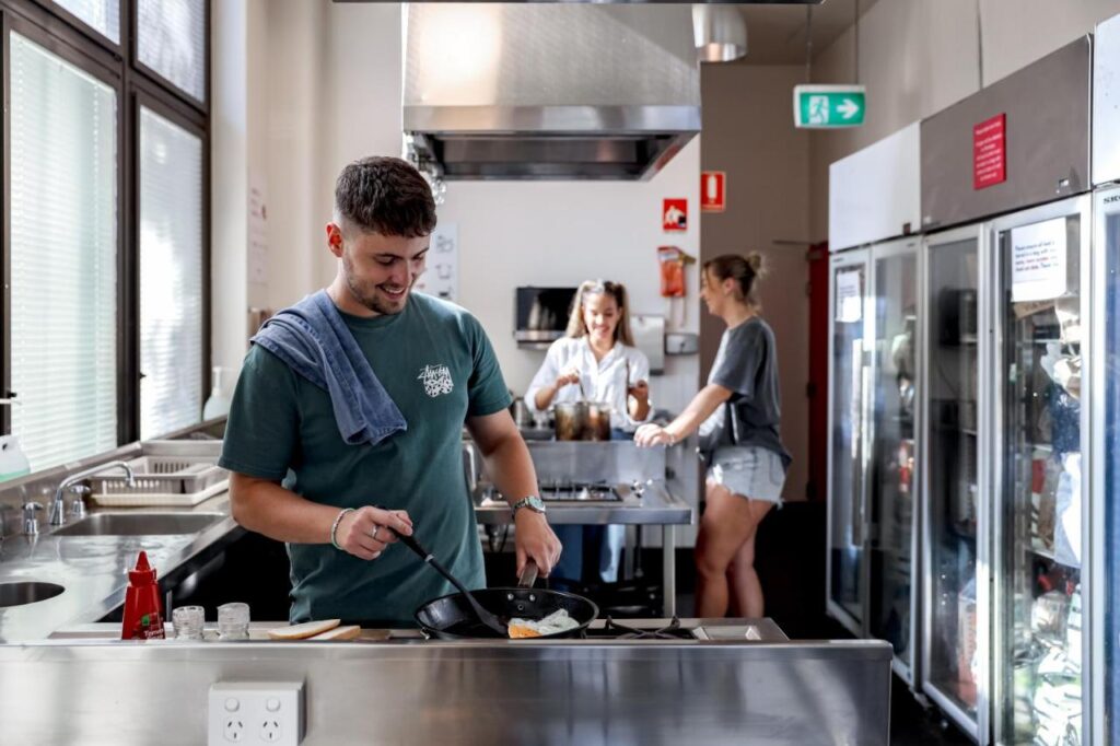 A traveller cooking egs while other travellers chat in the background in the kitchen at Hostel Wake Up! Sydney Central