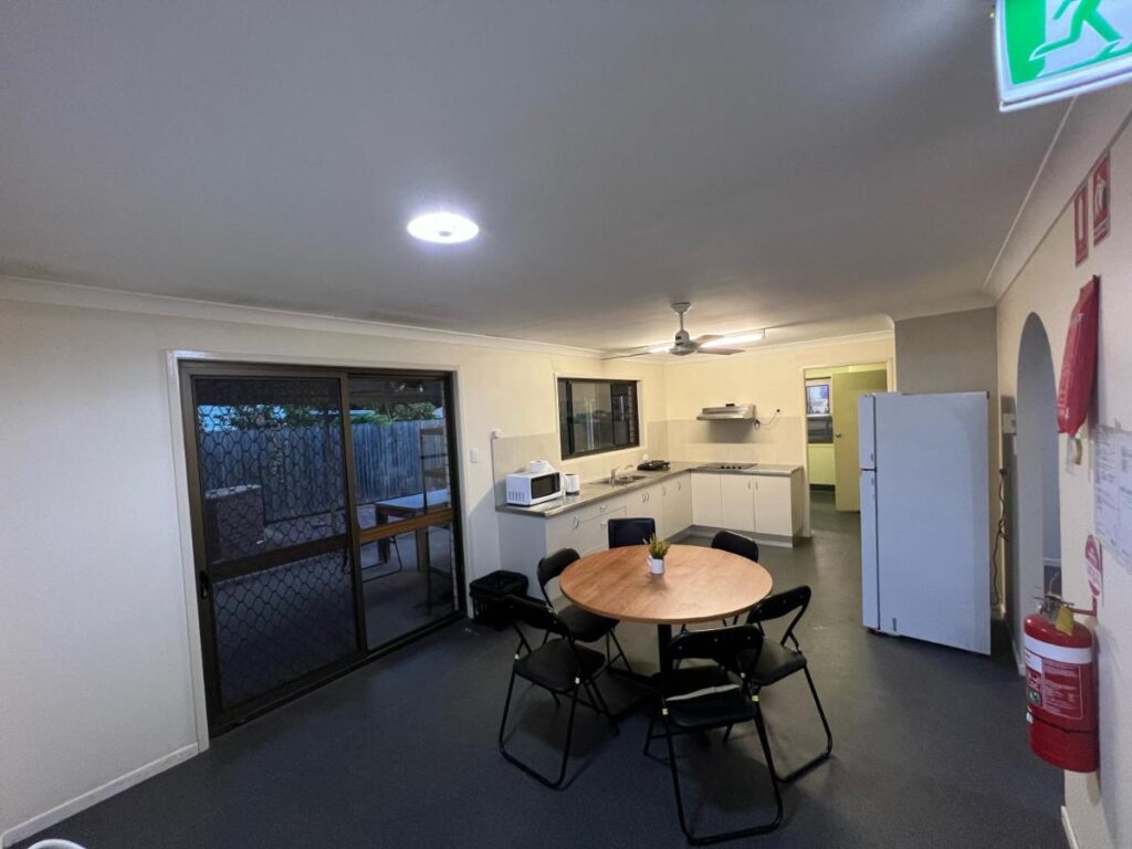 A shared kitchen space with a dining table, refrigerator, sink on an L-shaped bench top at Tomato Backpackers