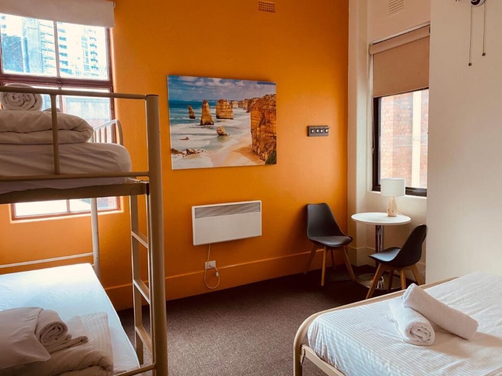 A three bed dormitory with a picture of te twelve apostles on a bright orange wall and a small table and chairs next to a window at YHA Melbourne Central